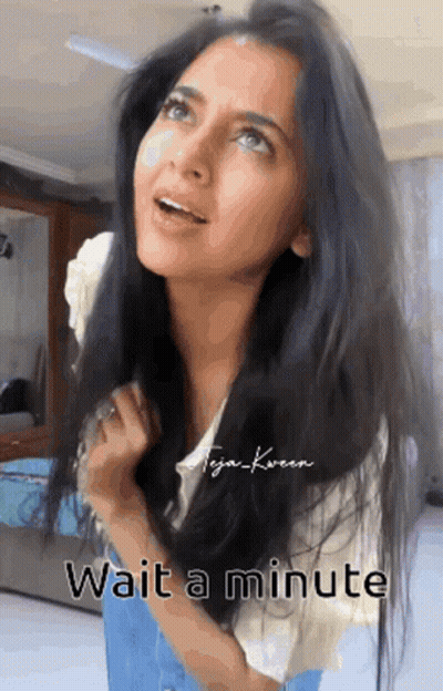 A Woman With Long Black Hair Is Standing In A Room With The Words Wait A Minute Below Her . GIF