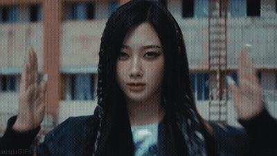 A Woman With Long Black Hair Is Standing In Front Of A Building With Her Hands In The Air GIF