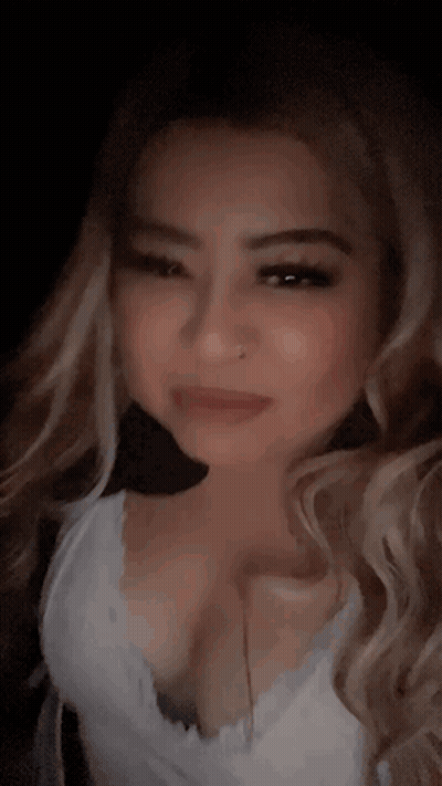 A Woman With Long Blonde Hair And A Nose Ring Is Wearing A White Tank Top And Looking At The Camera . GIF