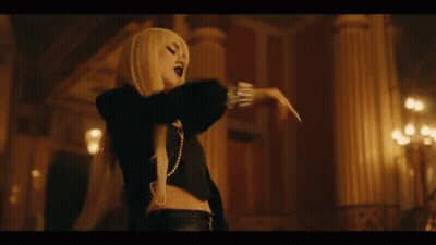 A Woman With Long Blonde Hair And A Pearl Necklace Is Standing In A Dark Room With Her Arms Outstretched . GIF