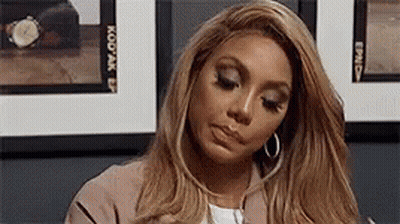 A Woman With Long Blonde Hair And Hoop Earrings Is Sitting In Front Of A Wall Of Pictures . GIF