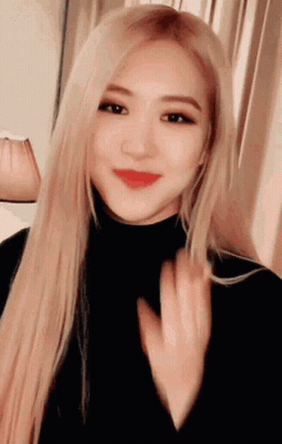 A Woman With Long Blonde Hair And Red Lips Is Wearing A Black Turtleneck And Waving . GIF