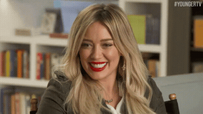 A Woman With Long Blonde Hair And Red Lipstick Is Smiling While Sitting In A Chair . GIF