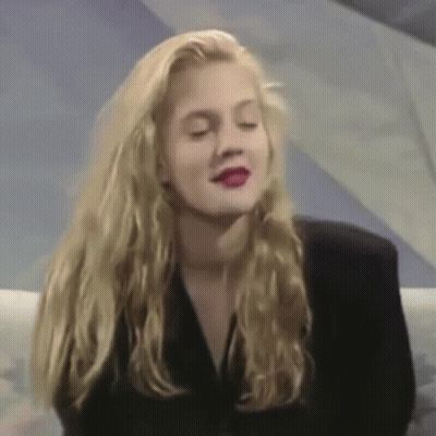 A Woman With Long Blonde Hair And Red Lipstick Is Wearing A Black Jacket And Sitting On A Couch . GIF