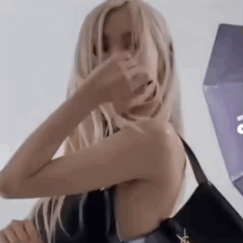 A Woman With Long Blonde Hair Is Holding A Black Bag And Covering Her Face With Her Hand . GIF