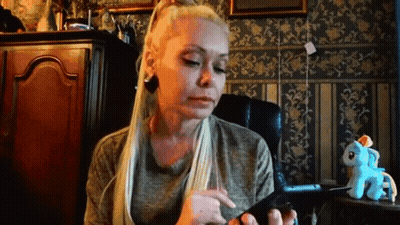 A Woman With Long Blonde Hair Is Sitting In A Chair Looking At Her Phone . GIF