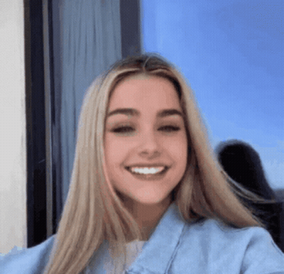A Woman With Long Blonde Hair Is Smiling For The Camera While Wearing A Blue Denim Jacket . GIF