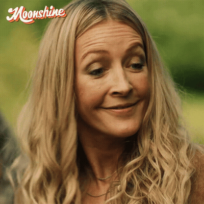 A Woman With Long Blonde Hair Is Smiling With A Moonshine Logo Behind Her GIF