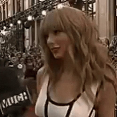 A Woman With Long Blonde Hair Is Standing In Front Of A Building In A White Tank Top . GIF