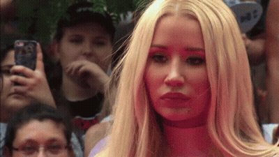 A Woman With Long Blonde Hair Is Standing In Front Of A Crowd While A Man Takes A Picture With His Phone GIF