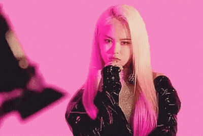 A Woman With Long Blonde Hair Is Standing In Front Of A Pink Background With Her Hands On Her Chin . GIF