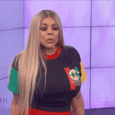 A Woman With Long Blonde Hair Is Standing In Front Of A Wall Wearing A Colorful Shirt . GIF