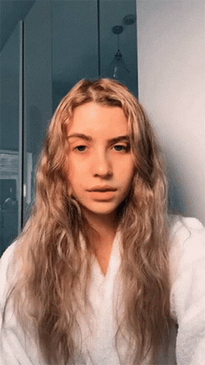 A Woman With Long Blonde Hair Is Wearing A White Robe And Taking A Selfie GIF