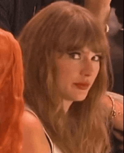 A Woman With Long Brown Hair And Bangs Is Sitting In A Crowd Of People And Looking At The Camera . GIF