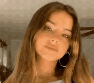 A Woman With Long Brown Hair And Hoop Earrings Is Looking At The Camera In A Living Room . GIF