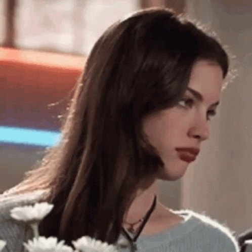 A Woman With Long Brown Hair Is Holding A Bouquet Of Daisies And Looking At The Camera . GIF