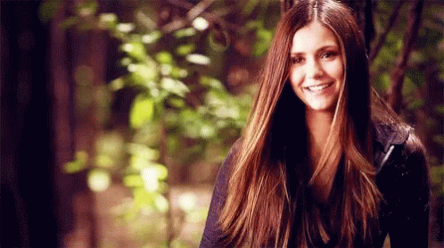 A Woman With Long Brown Hair Is Smiling While Standing In Front Of Trees . GIF
