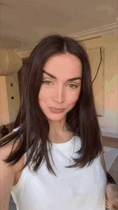 A Woman With Long Brown Hair Is Wearing A White Tank Top And Necklace . GIF