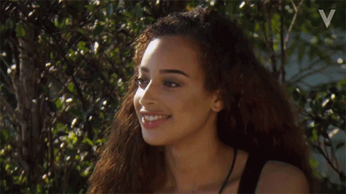 A Woman With Long Curly Hair Is Smiling While Standing In Front Of A Tree . GIF