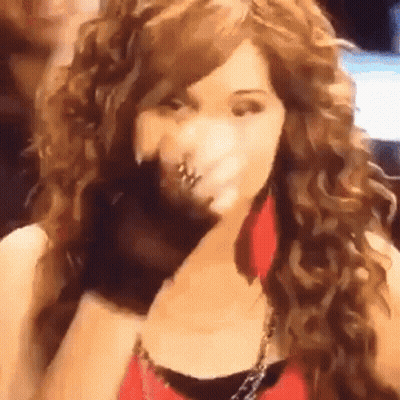 A Woman With Long Curly Hair Is Wearing A Red Top And Earrings And Covering Her Face With Her Hand . GIF