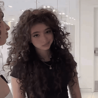A Woman With Long Curly Hair Wearing A Black Shirt GIF