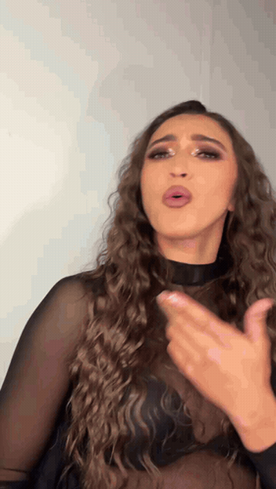 A Woman With Long Curly Hair Wearing A Black Top Is Making A Funny Face . GIF