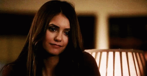 A Woman With Long Dark Hair Is Sitting In Front Of A Lamp And Looking At The Camera . GIF