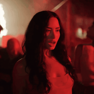 A Woman With Long Dark Hair Is Standing In A Dark Room With Red Lights Behind Her GIF
