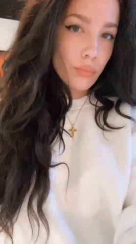 A Woman With Long Dark Hair Is Wearing A White Shirt And A Cross Necklace . GIF