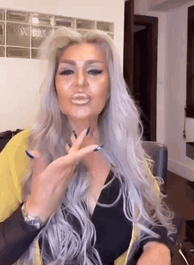 A Woman With Long Gray Hair And Blue Nails Is Sitting At A Table Making A Funny Face . GIF