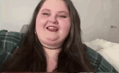 A Woman With Long Hair And A Choker Is Sitting On A Bed And Smiling . GIF