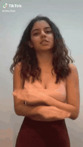 A Woman With Long Hair And A Very Large Breast Is Standing With Her Arms Crossed In Front Of A White Wall . GIF