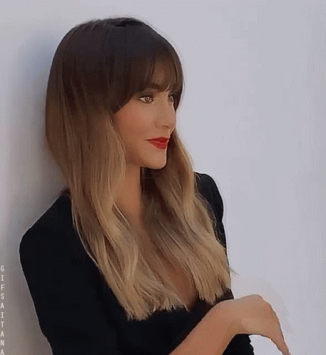 A Woman With Long Hair And Bangs Is Leaning Against A Wall . GIF