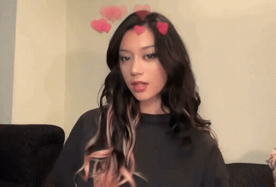 A Woman With Long Hair And Pink Highlights Is Sitting On A Couch With Hearts On Her Head . GIF