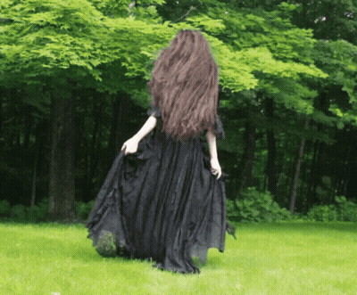 A Woman With Long Hair In A Black Dress Stands In The Grass GIF