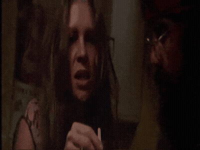 A Woman With Long Hair Is Holding A Cigarette And Talking To A Man With Glasses GIF