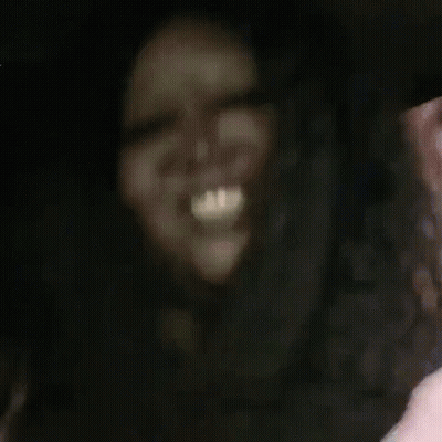 A Woman With Long Hair Is Laughing And Waving At The Camera While Sitting In A Car . GIF