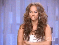 A Woman With Long Hair Is Sitting In A Chair With Her Hands Folded In Front Of Her Face . GIF