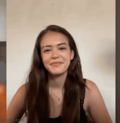 A Woman With Long Hair Is Smiling And Looking At The Camera While Sitting In Front Of A Wall . GIF