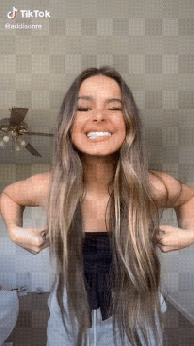 A Woman With Long Hair Is Smiling While Standing In Front Of A Ceiling Fan . GIF