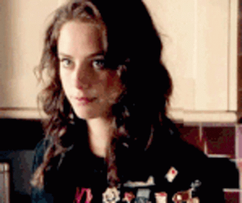 A Woman With Long Hair Is Standing In A Kitchen Wearing A Black Sweater With Stickers On It . GIF