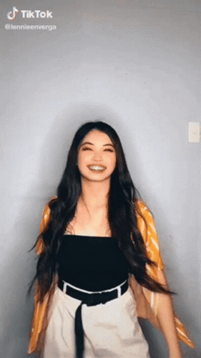 A Woman With Long Hair Is Wearing A Black Top And White Shorts And A Yellow Cardigan . GIF