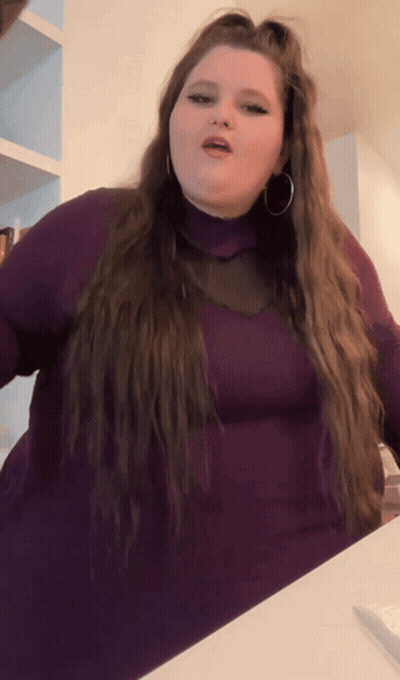 A Woman With Long Hair Is Wearing A Purple Turtleneck And Hoop Earrings GIF