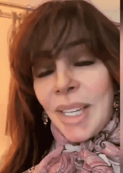 A Woman With Long Hair Is Wearing A Scarf Around Her Neck And Smiling . GIF