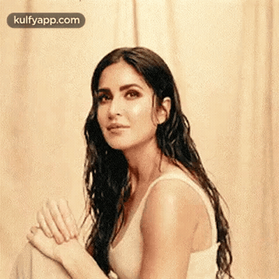 A Woman With Long Hair Is Wearing A White Tank Top And Sitting In Front Of A Curtain . GIF