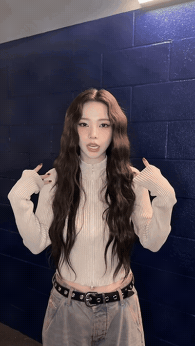 A Woman With Long Hair Wearing A White Sweater And Jeans Stands In Front Of A Blue Brick Wall GIF