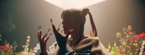 A Woman With Long Nails And A Ponytail Is Standing In Front Of Flowers . GIF
