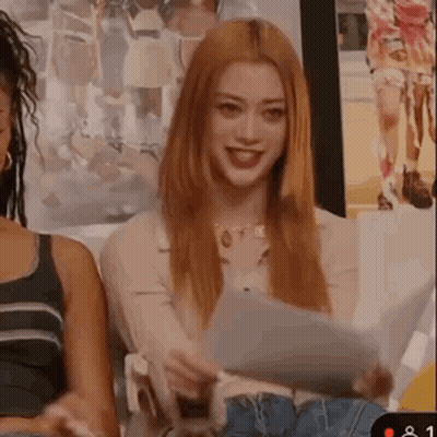 A Woman With Long Red Hair Is Holding A Piece Of Paper And Smiling While Sitting In Front Of A Group Of People . GIF