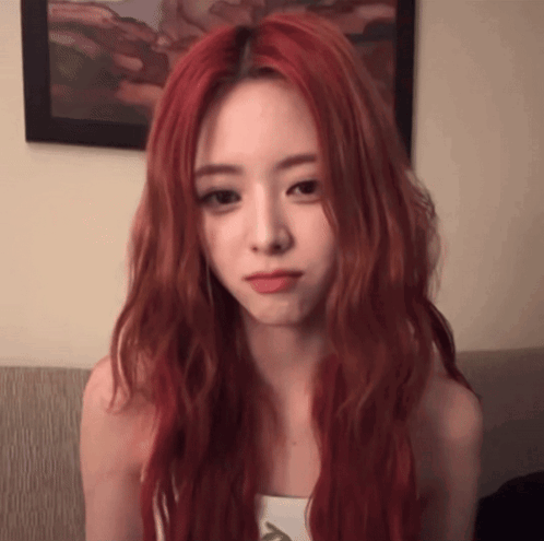 A Woman With Long Red Hair Is Sitting On A Couch And Making A Face . GIF