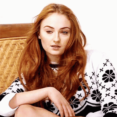 A Woman With Long Red Hair Is Sitting On A Wicker Chair Wearing A Black And White Sweater With Flowers On It . GIF